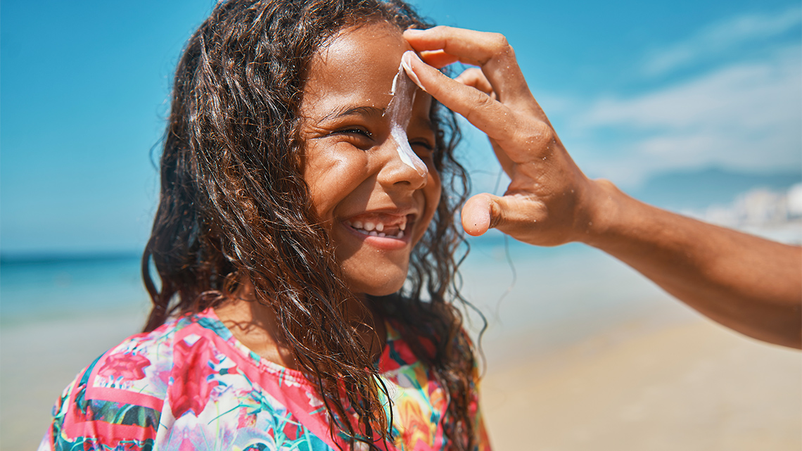 An adult’s hand applies sunscreen to a child’s face on a beach.