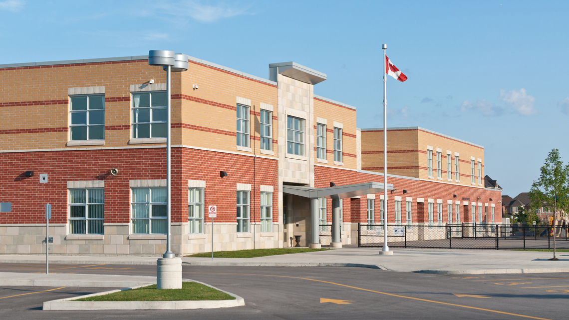 A 2-storey brick building with many windows next to a parking lot and a fenced-in playground.