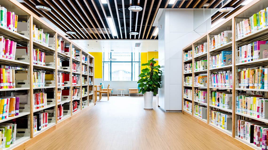 A pathway with two large bookshelves one on each side. There are table and chairs in the background.