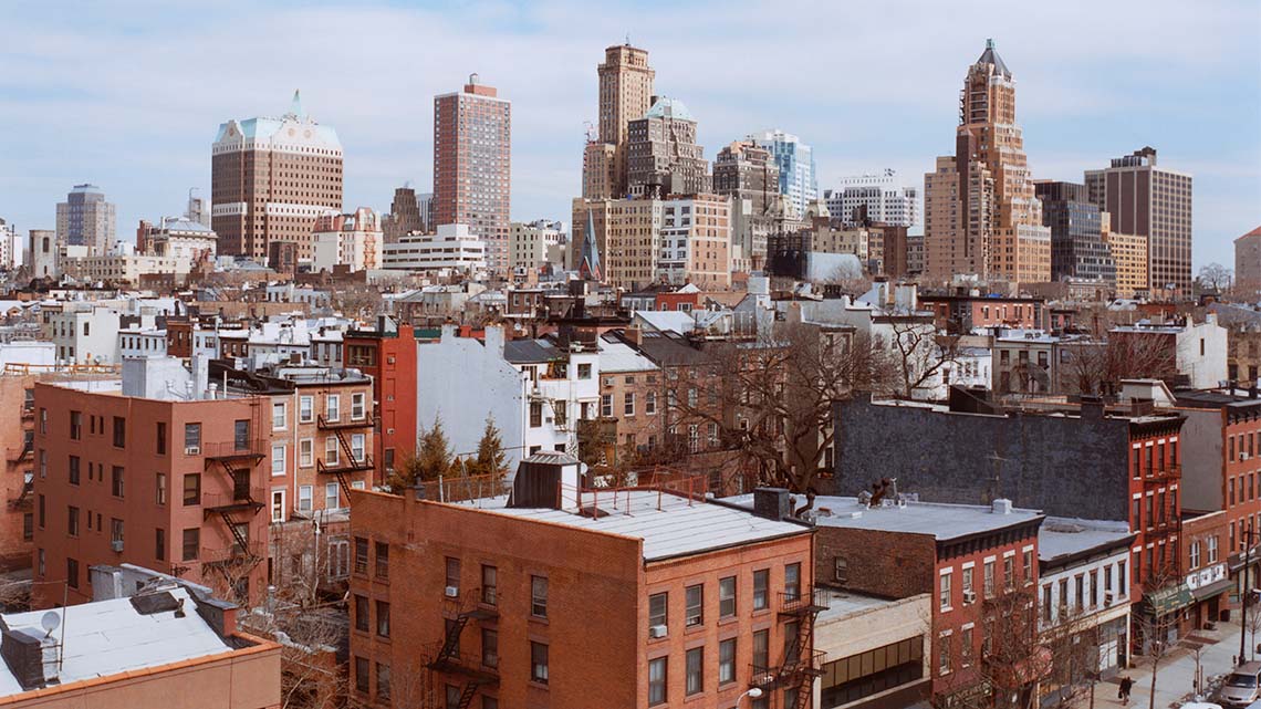 This is an image of a cityscape. There are tall buildings and shorter buildings. The windows of many of the buildings are the same. There are cars parked on the road in front of the buildings.