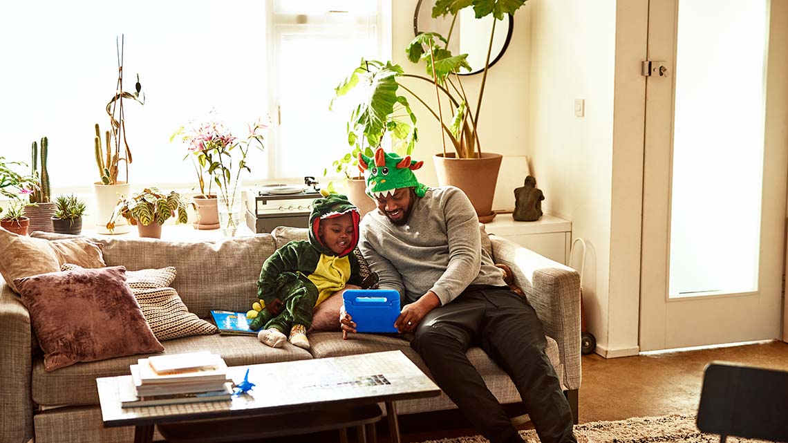 A child and an adult use a device. There are plants on a table behind the couch. There is a table in front of them and door beside them.