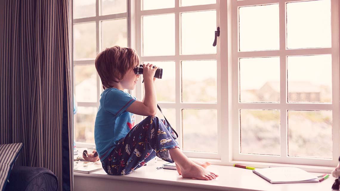 A child is looking out their window with binoculars to view the outside world. They have pens and notebooks in the space around them.