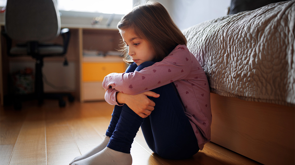A child is sitting on the floor frowning with their arms wrapped around their  legs.