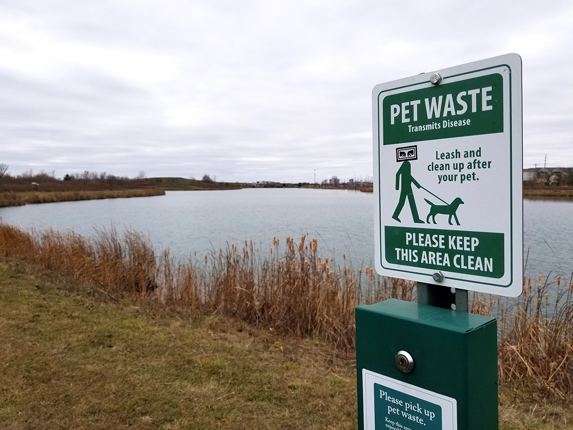 Sign placed in the ground in front of a dirty pond. The sign reads “Pet waste transmits disease. Please keep this area clean.