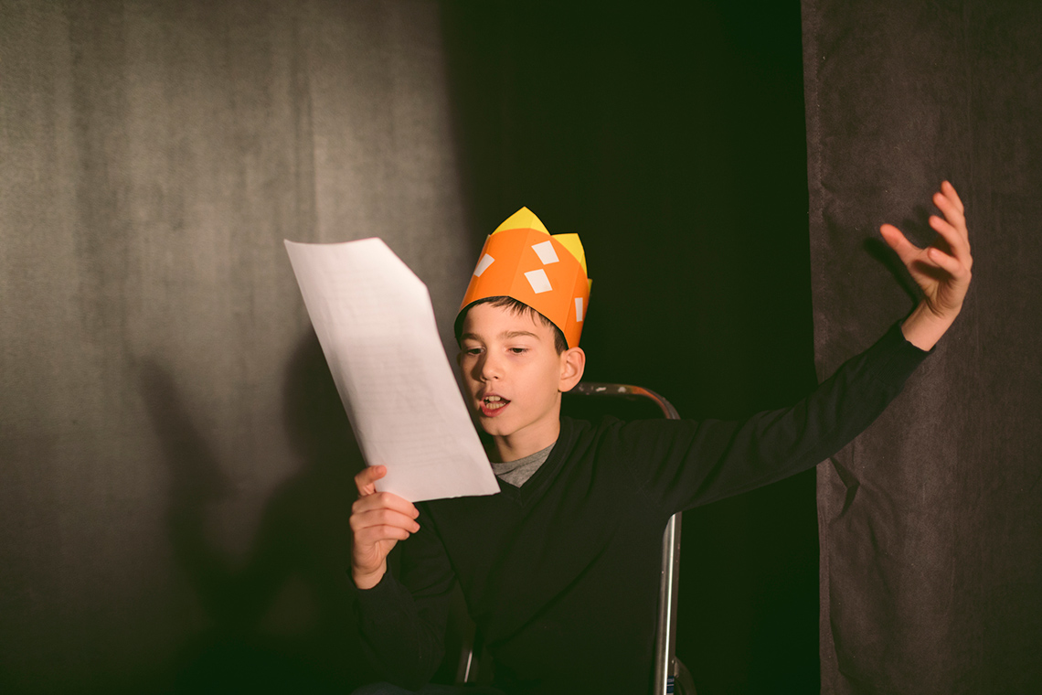 A young student reads lines from a script and makes a dramatic gesture with one of their hands.