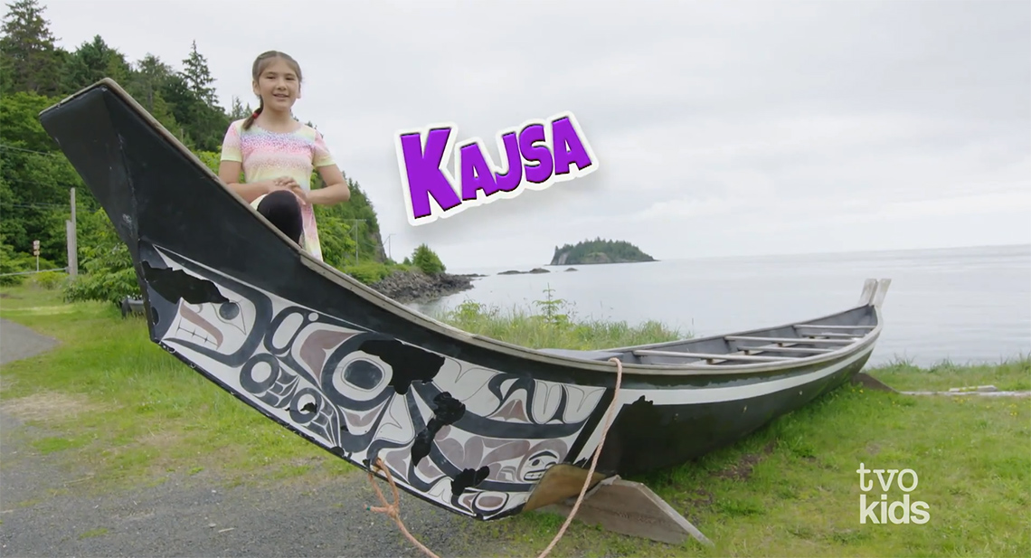 An image from Raven’s Quest: Kajsa video. Kajsa is standing by the beach with a canoe in front of her.