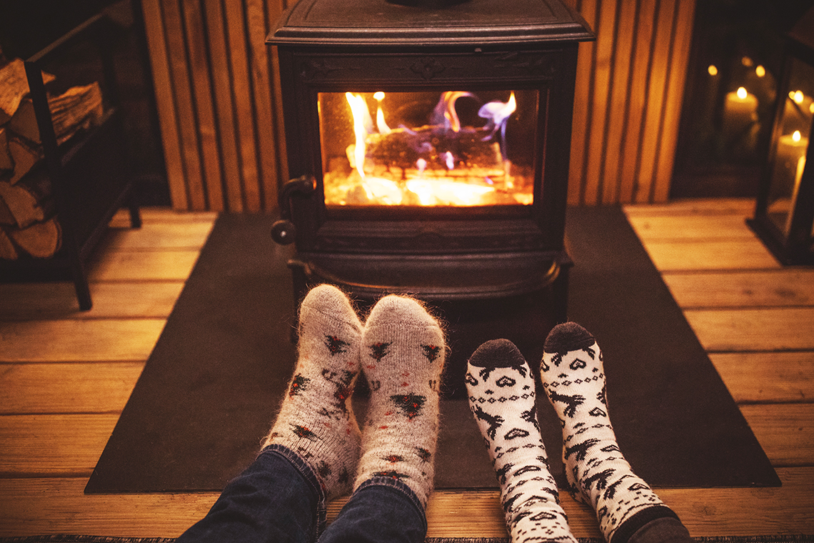 An image of a cozy fireplace. The image shows two people’s feel extended in front of the fireplace for warmth.
