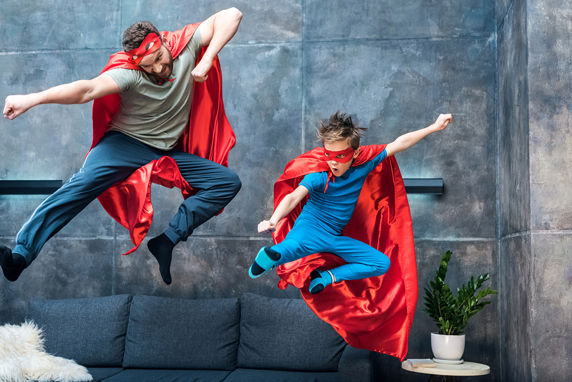 An adult and a child dressed like superheroes are running in a park with trees in the background.