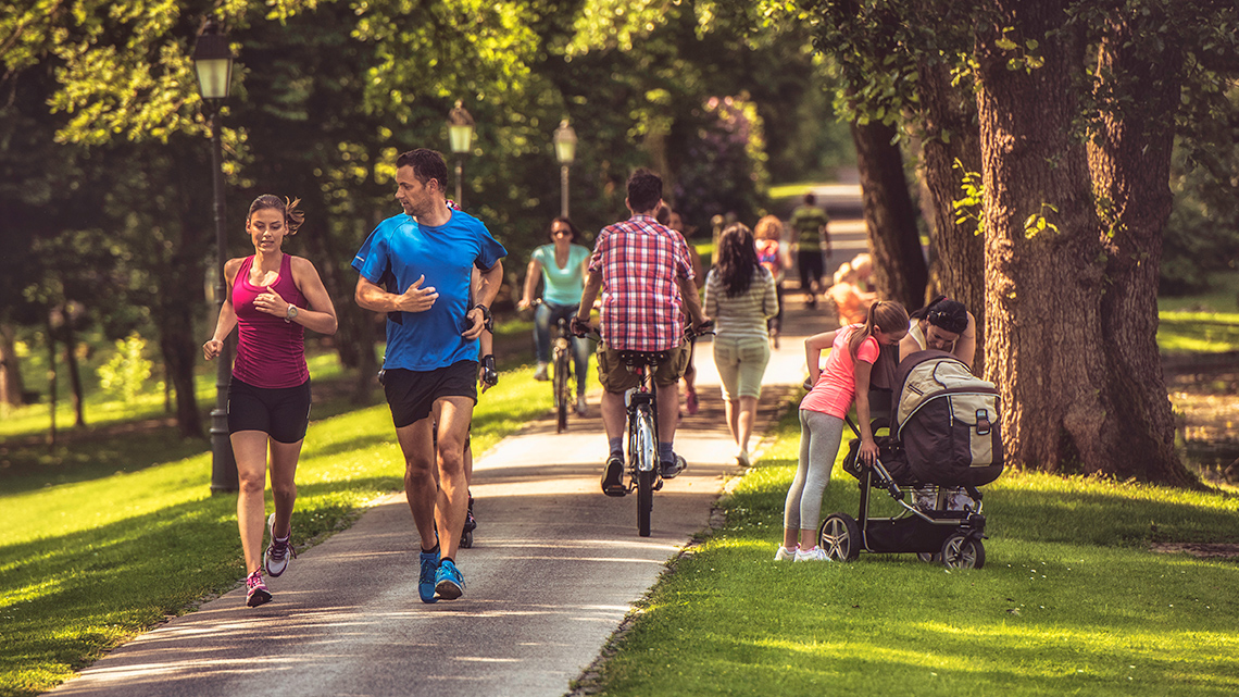 A woman and a man jogging in the park. A woman and a girl are looking in a baby carriage.