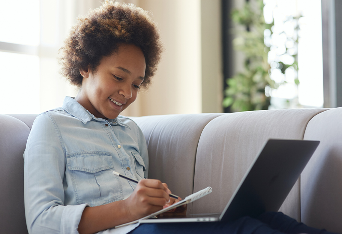 Student sitting on a sofa using a laptop and writing notes.