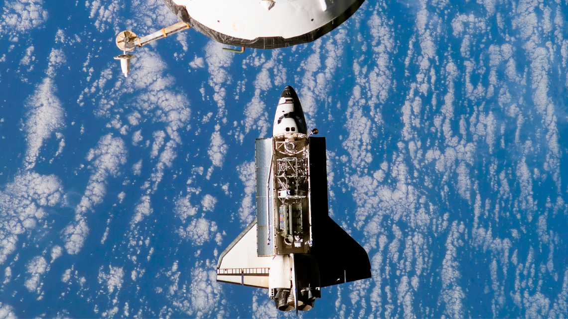 Astronauts work on a space station while a space shuttle approaches