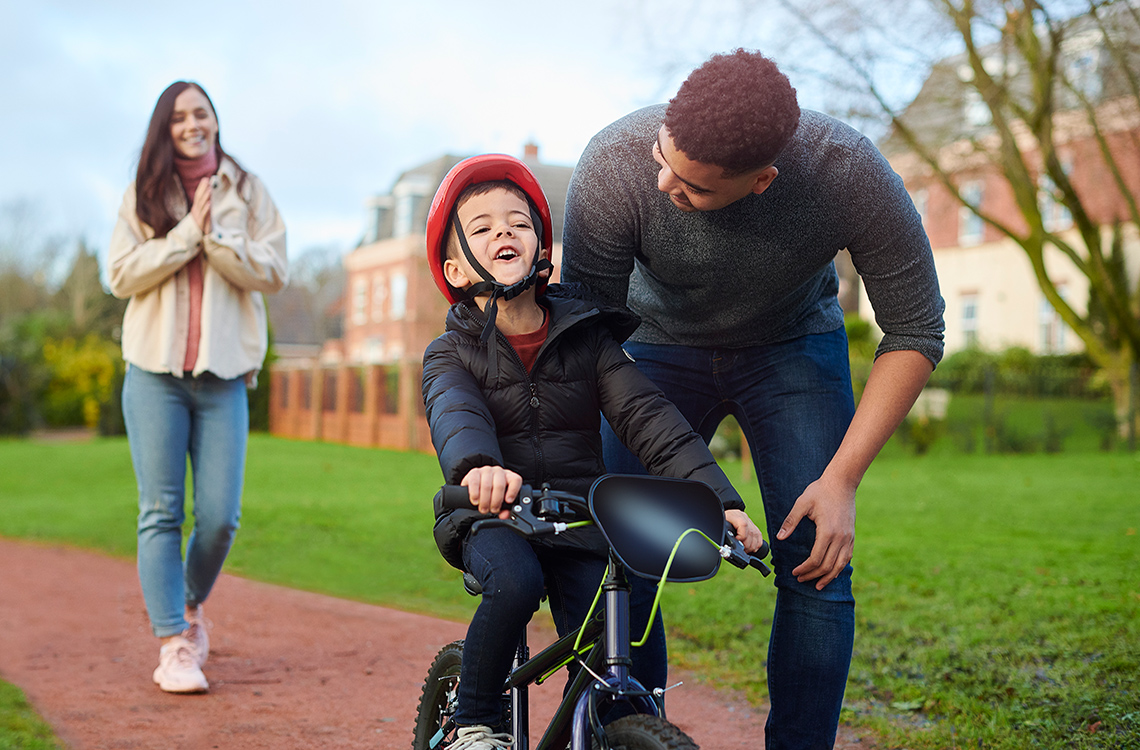 2 parents guide a child to ride a tricycle while wearing a helmet.