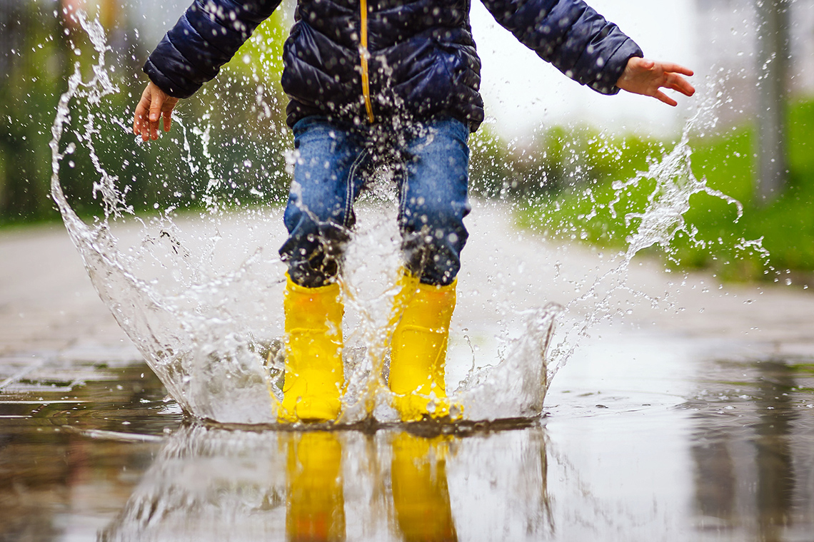Child with rain boots and umbrella jumping in a large puddle.