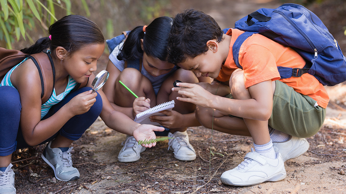 Three young students squat on the ground outside. The students on the left and right are using magnifying glasses to observe a leaf held out by the student on the left. The student in the middle is writing on a notepad.