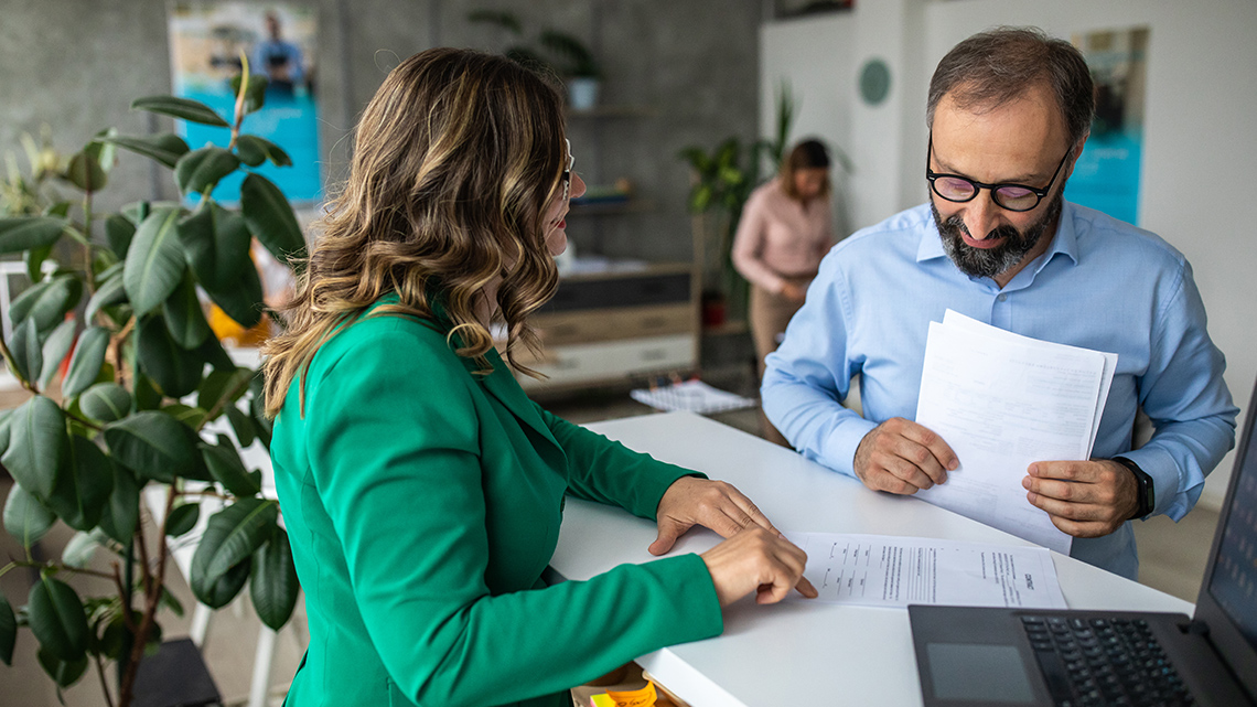 A person at a bank with some paperwork talking to a bank teller.