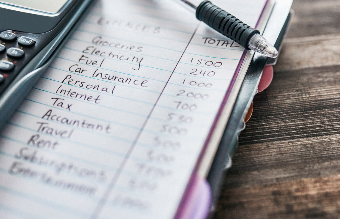 A notebook with a list of monthly expenses. On the notebook is a calculator and a pen.