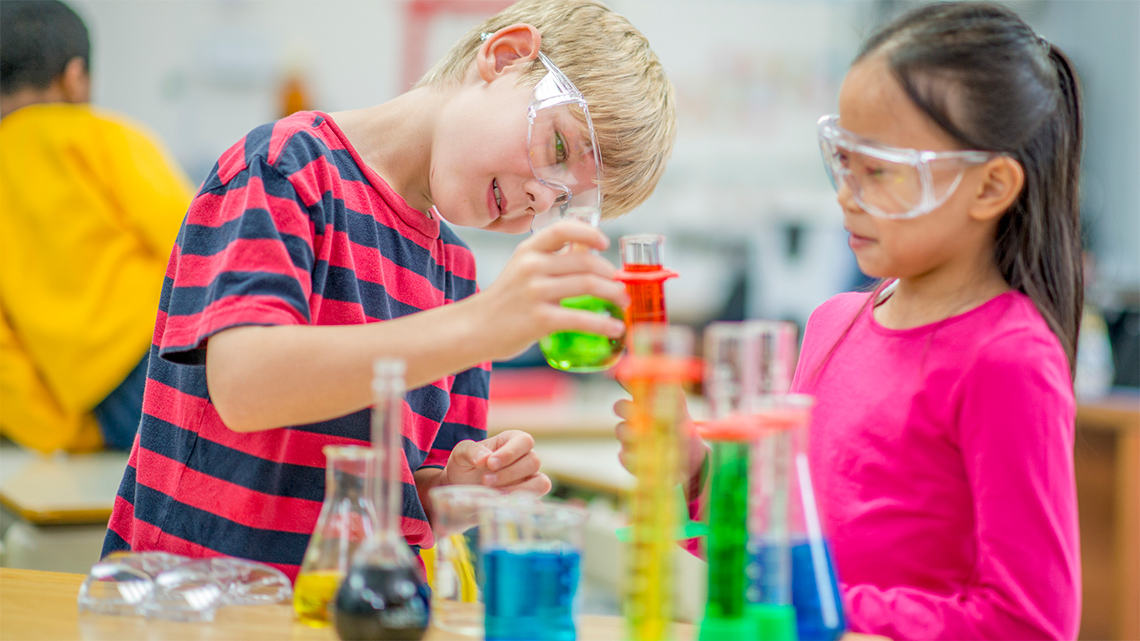 Students wearing protective glasses and pouring a liquid into the beaker.