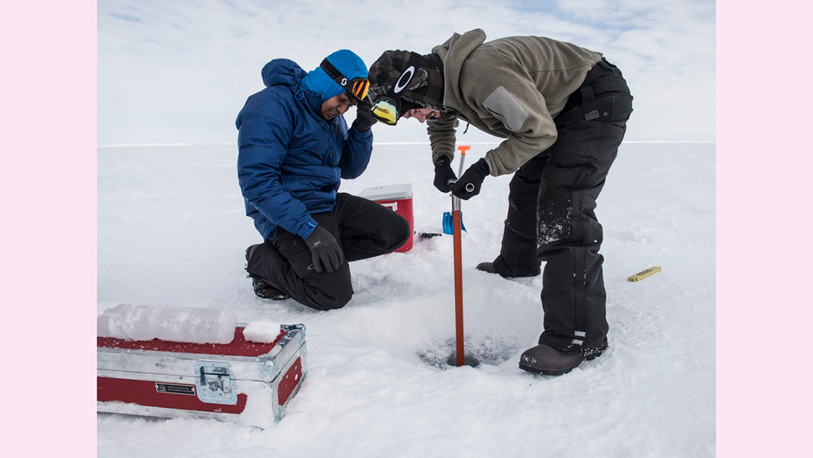 Two scientists are collecting ice samples using a tool called an auger. They are wearing ski googles, hats, coats, gloves, snowpants, and winter boots to keep warm.