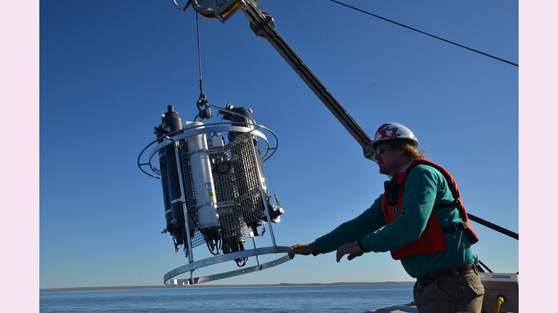 A scientist is using a crane and some tubes to do his research. He is wearing a helmet, sunglasses, a lifejacket, gloves, and a coat.
