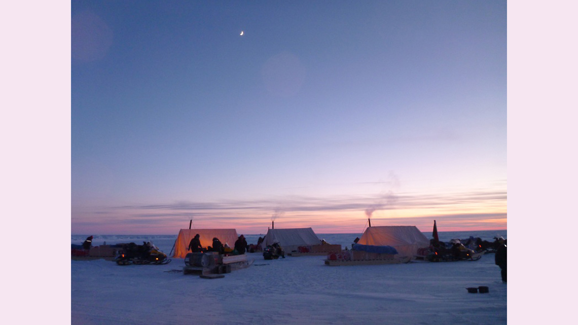 Scientists are keeping warm inside heated tents in the snow-covered Arctic at sunset. There is smoke coming from the tents.