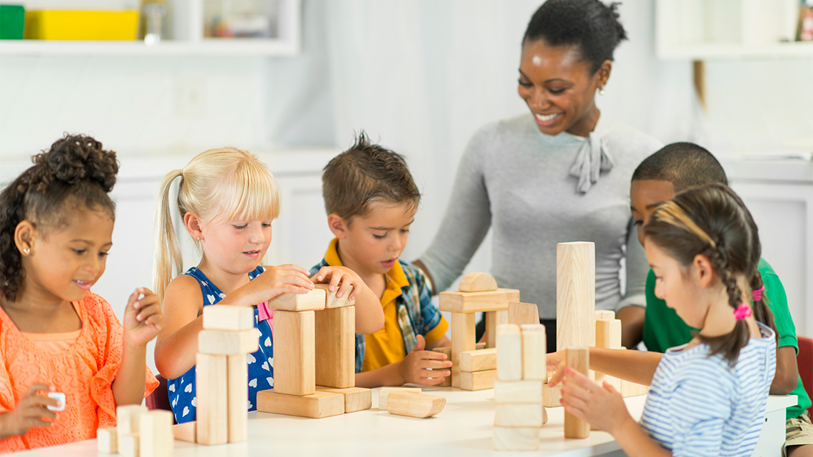 Five children and an adult build structures using wooden blocks. 