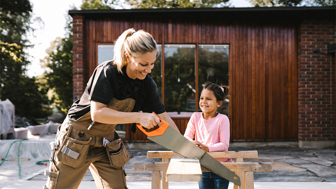 An adult cuts a piece of wood using a hand saw, while a child watches. 
