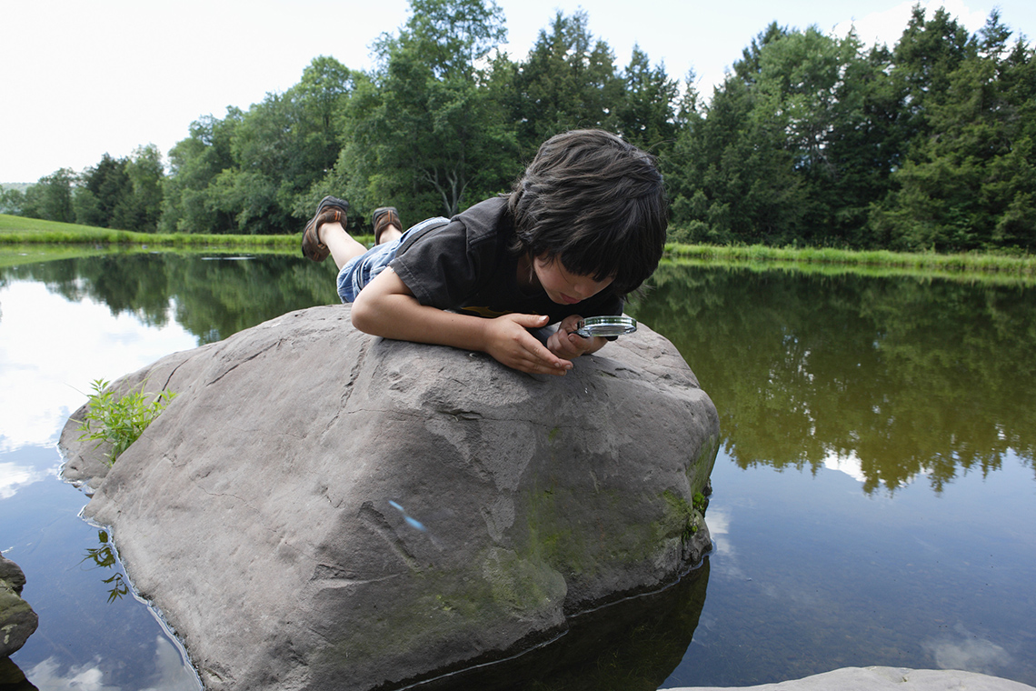 A young person lays on top of a rock, in the middle of a lake or pond, and looks at the water through a magnifying glass on a clear day.