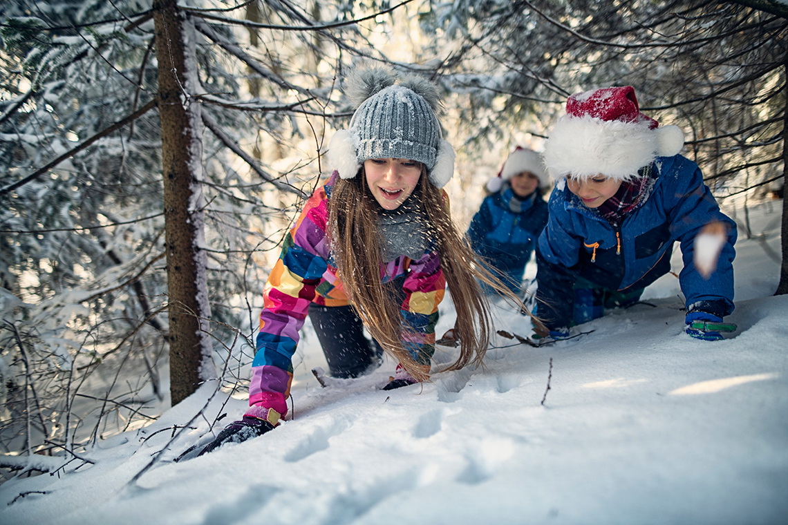 Three young people, dressed in winter clothing, crawl on their hands and knees underneath some trees, as they follow tracks in the snow.