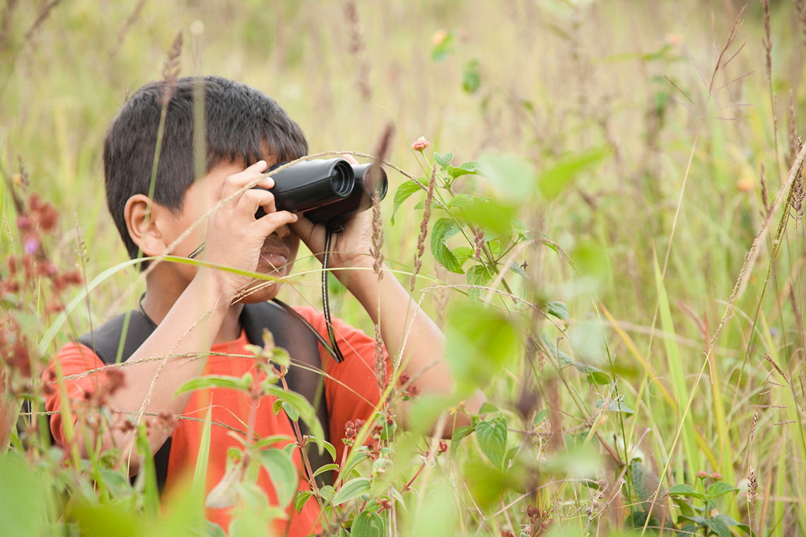 A young person wearing a backpack stands in tall grasses and looks through a pair of binoculars on a clear summer day.
