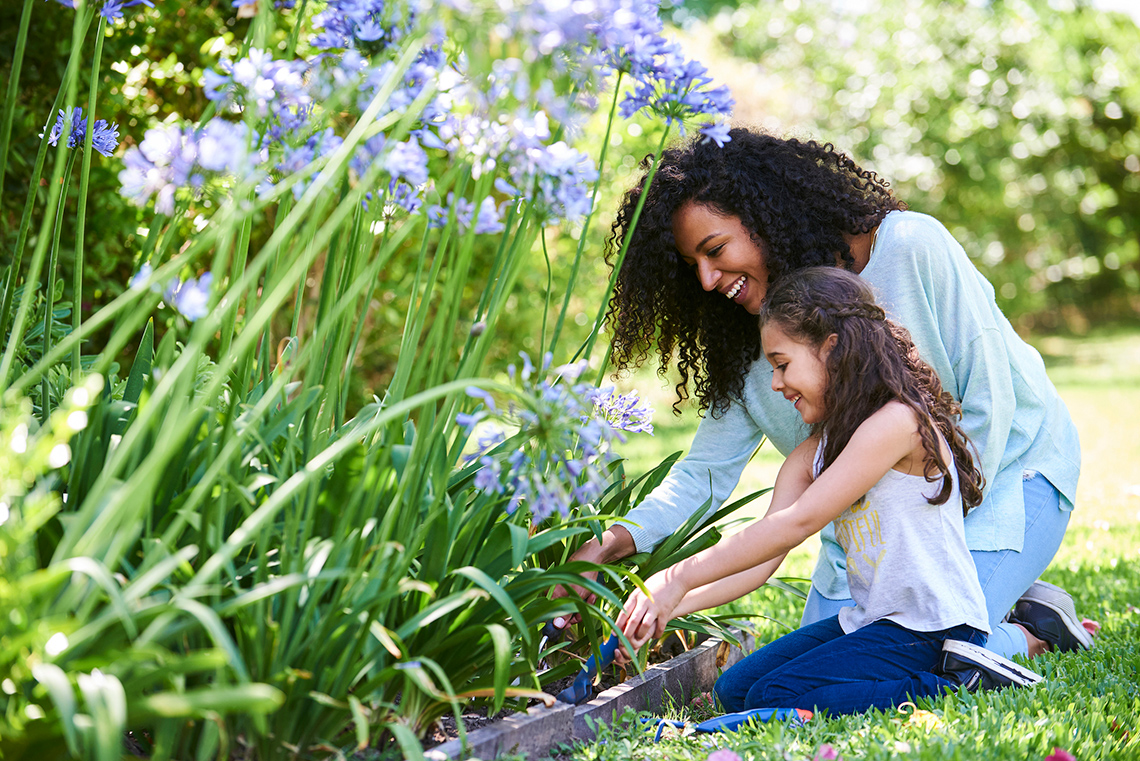 A woman and a young child kneel in front of a garden filled with milkweed and other plants, tending the soil with gardening tools.