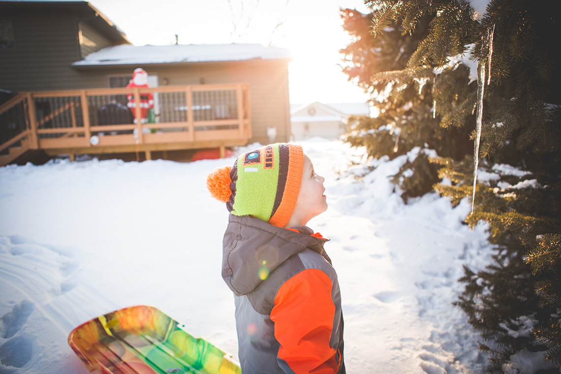 A young person pulling a sled stops to look at an icicle on a tree on a sunny winter day, a house with a Santa on a deck in the background.