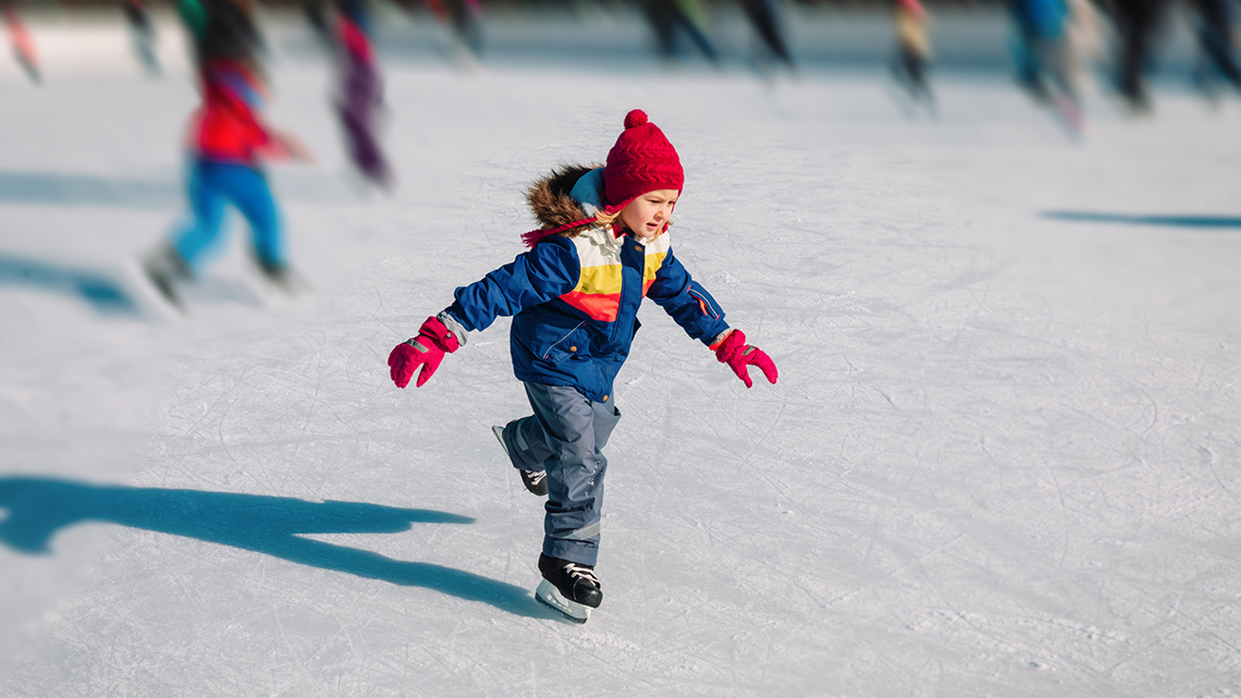 A young child wearing a hat, gloves, winter coat and snow pants holds their arms out as they skate on an outdoor ice rink on a sunny day.