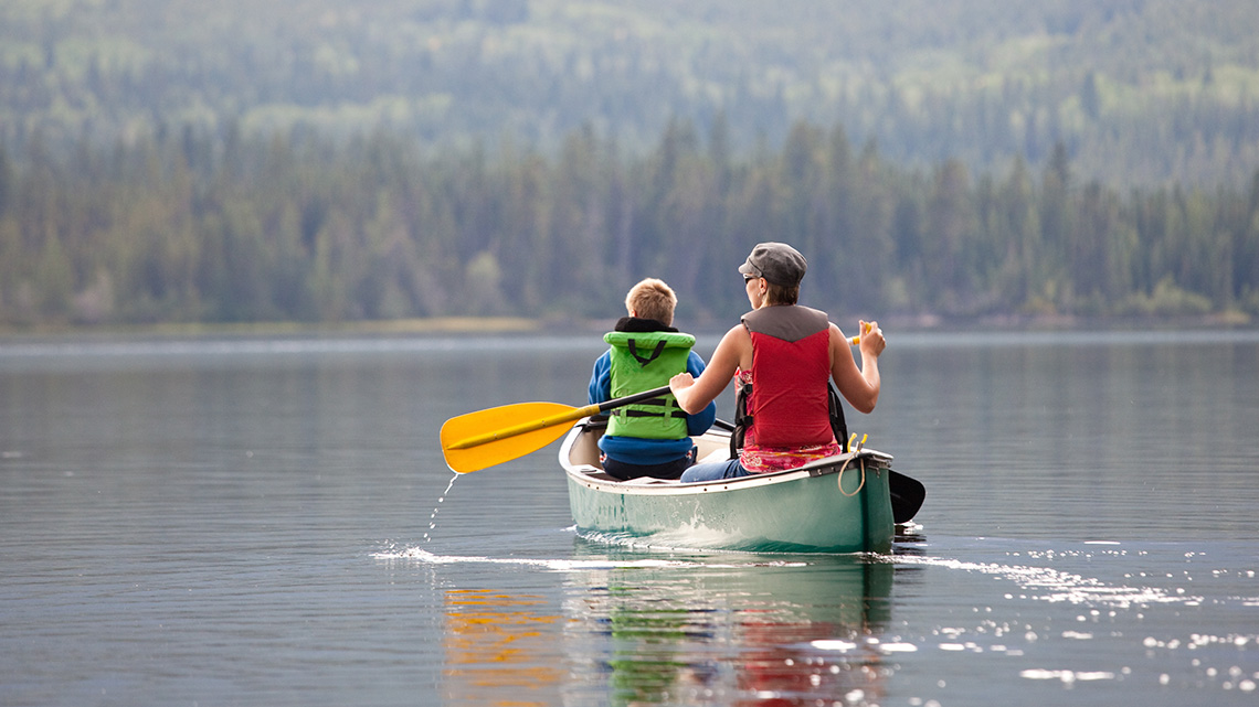 A guardian and child wearing lifejackets paddling a canoe in a lake.