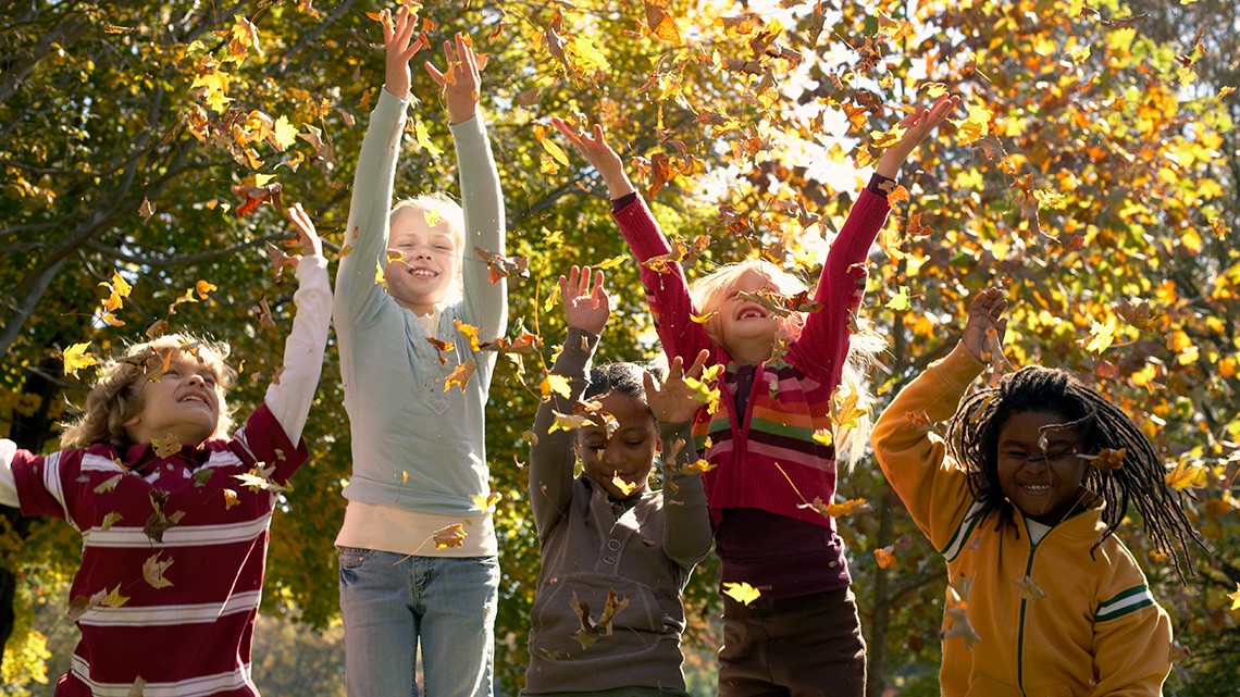 Children throwing leaves above their heads and jumping; the leaves are falling back down to the ground.