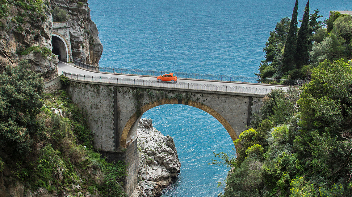 A car driving over an arch bridge above a river.