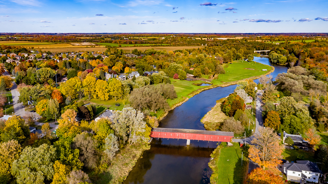 A bridge crosses a river flowing through a forested area containing several houses with farmland in the background.