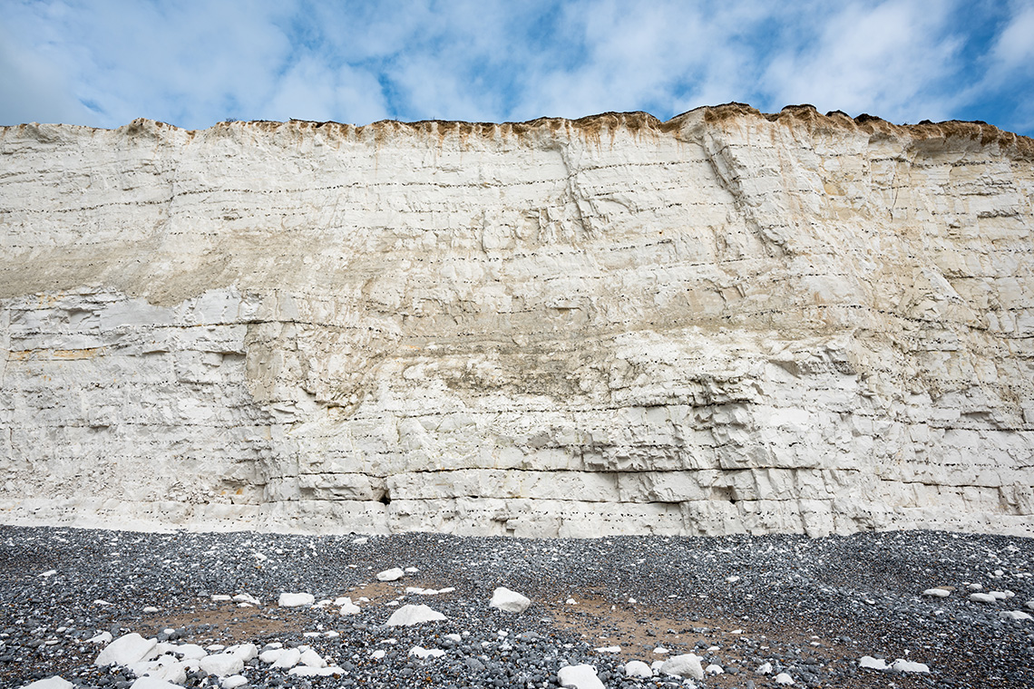Exposed, chalk-coloured rockface on the side of a cliff with a blue sky and clouds in the background.