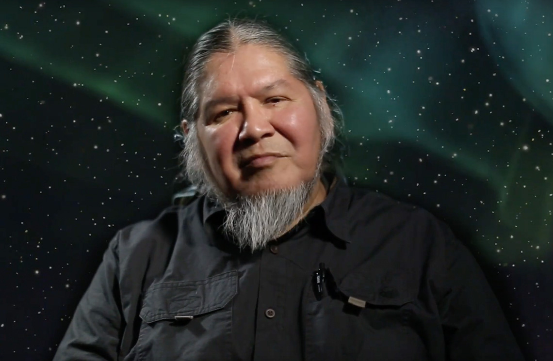 A middle-aged person with a white beard and his hair is pulled back is looking at the camera with a small smile on his face.
