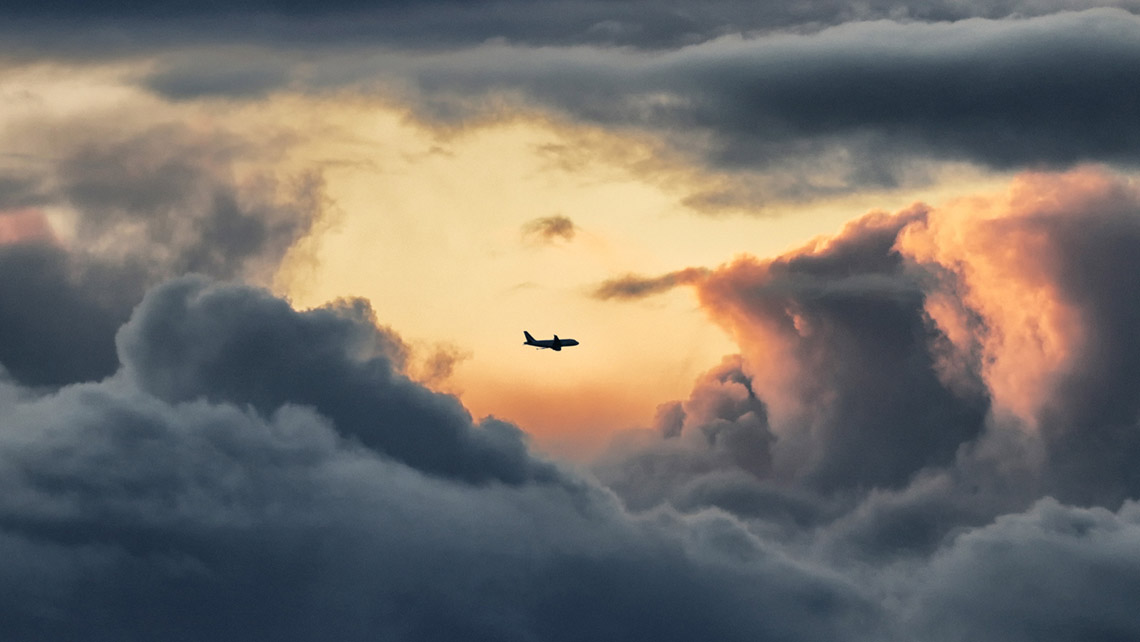 Dark clouds encircle a sunlit orange area of the sky. An airplane flies through the lit orange area.