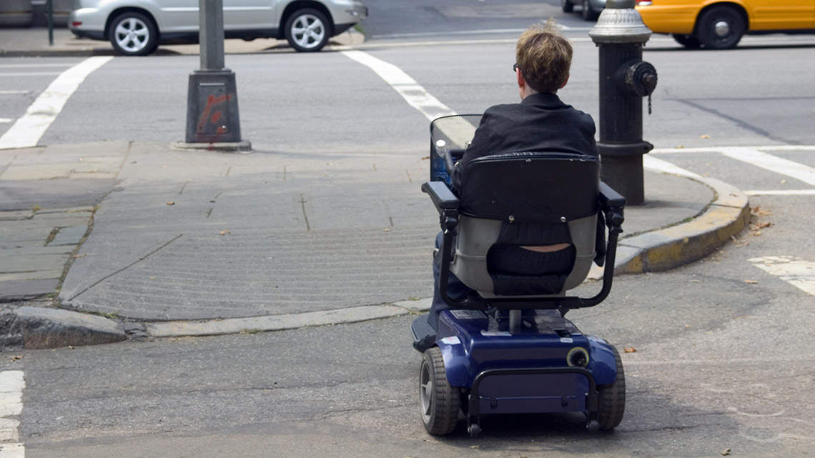 A person sits in a motorized scooter. They drive it across the street.