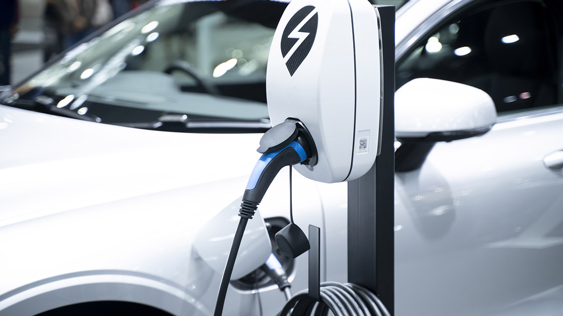 There is car that parked beside a post with a charging station. There is a heavy cord that is plugged into both the car and the charging station.