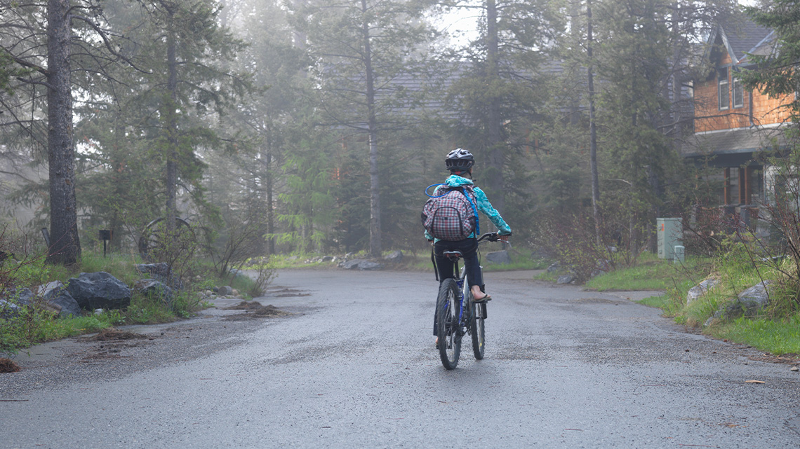 A child is biking on a road beside a home that has trees around it. The child has a backpack on their back and a helmet on their head.