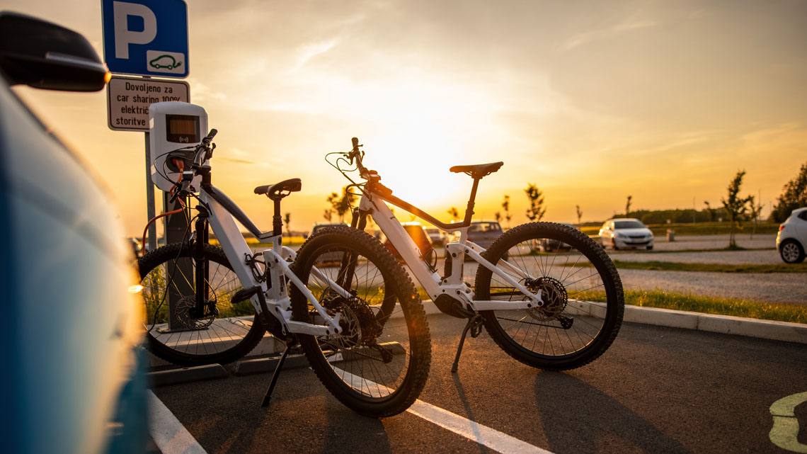 There are two electric bikes at a charging station beside a parking lot. The bikes are on kick stands and are plugged in to charge.