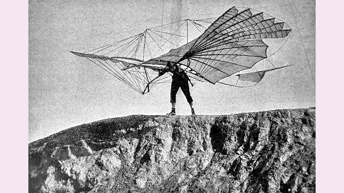 Historical photo of a person on a clifftop with a glider machine attached to their back.
