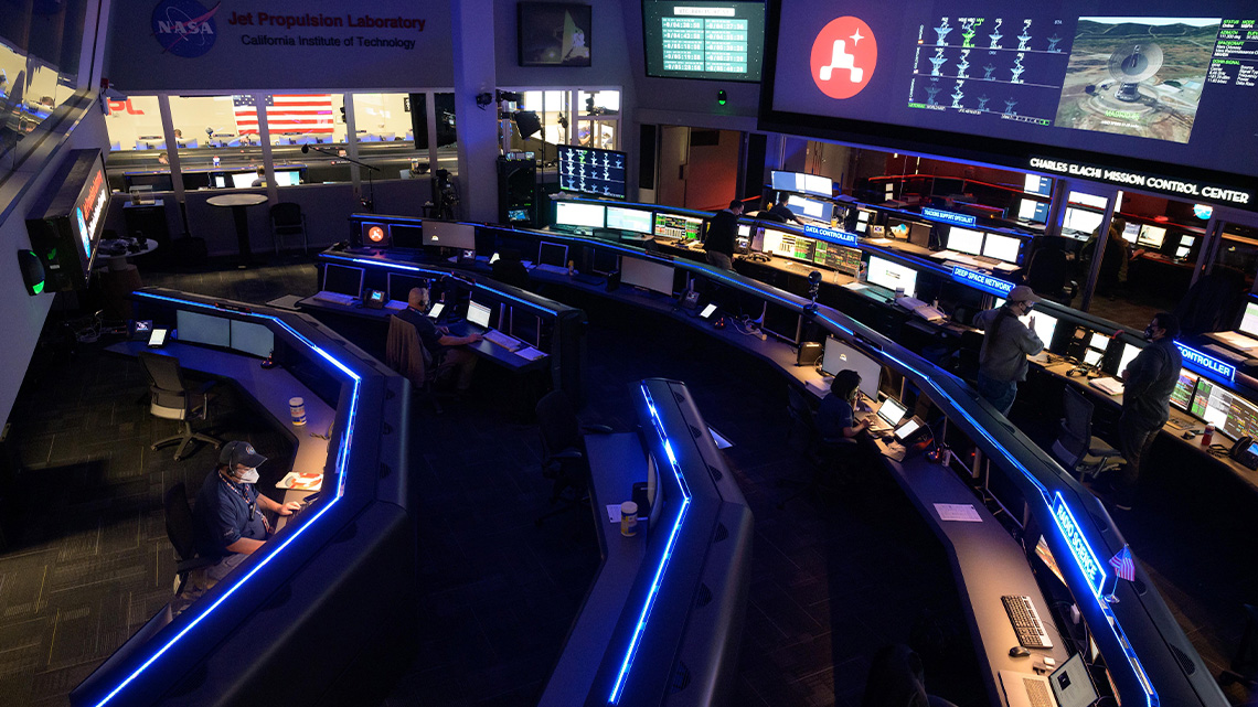 Large mission control room at NASA with many long desks and large screens. People are working on computers.