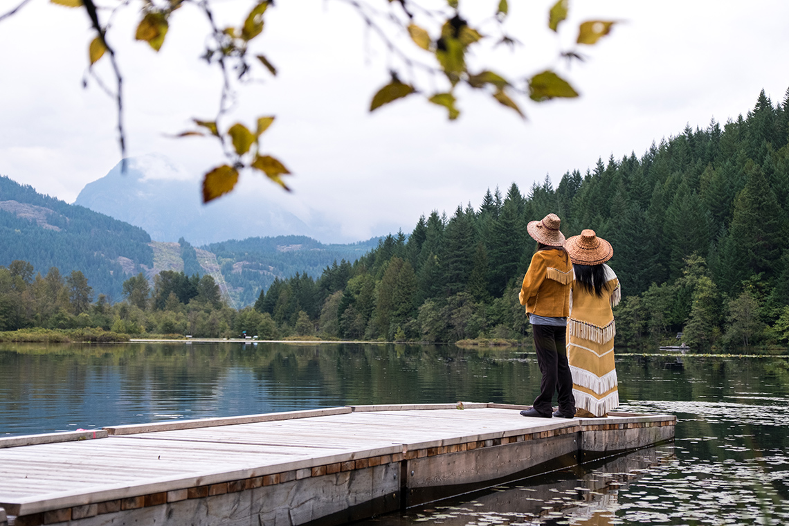 Two people standing on the edge of a dock looking out on to the water and surrounding mountains and forest.