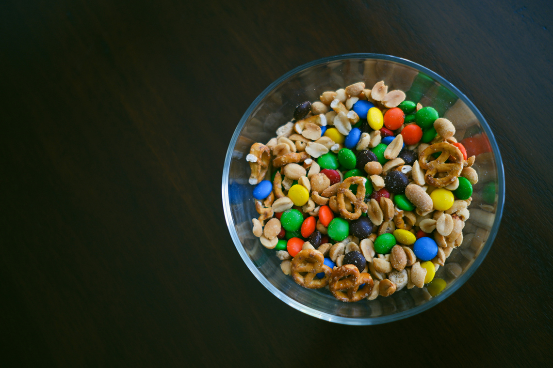A clear bowl containing peanuts, pretzels and candy-coated chocolate, on a black background
