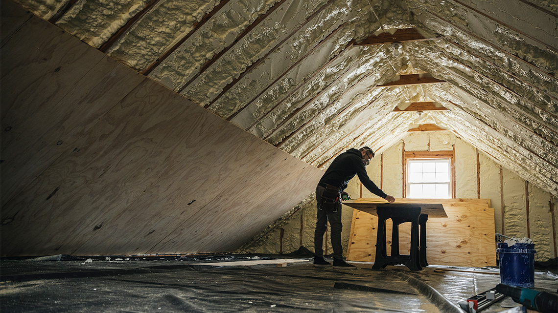 Person with safety glass and respirator mask spraying foam insulation on plywood boards in an attic.