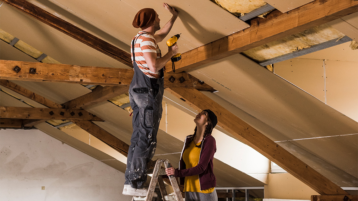 Two people attaching sheetrock to a slanted ceiling with insulation on it with a ladder and drill.