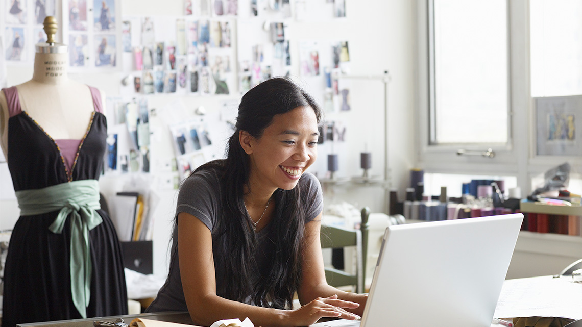 There is a young adult at their computer with a dress in the background on a mannequin.
